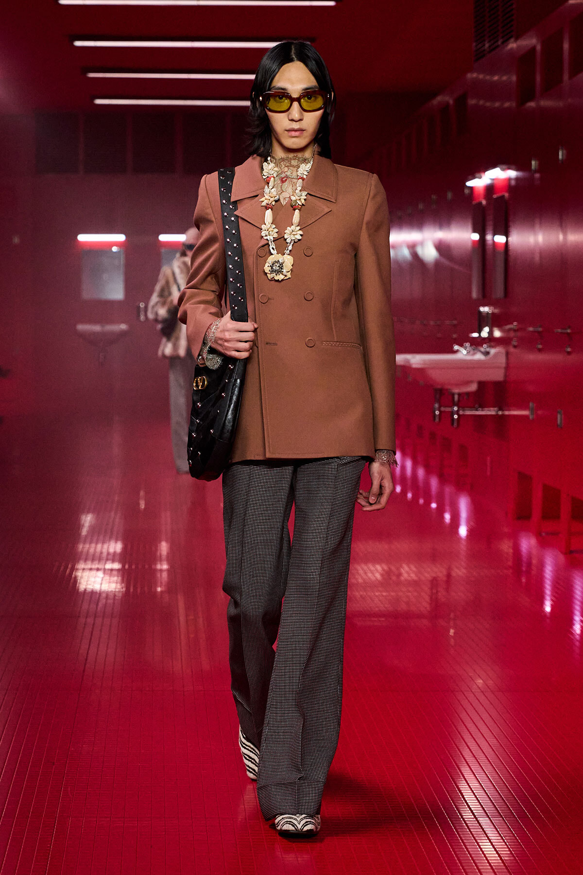 Model wearing a brown double-breasted blazer with gray plaid wide-leg pants, yellow sunglasses, and a large floral necklace, carrying a black studded bag in a red-lit hallway.