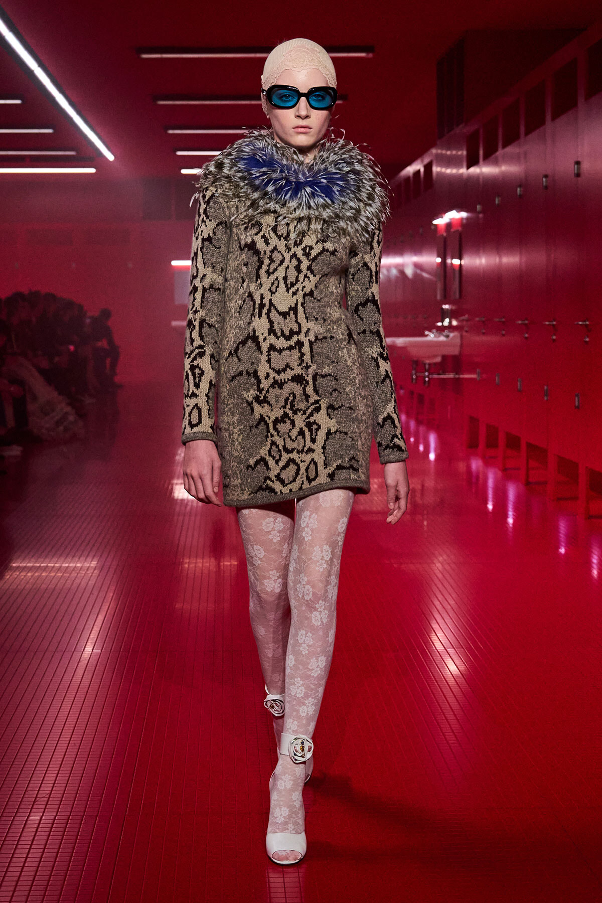 Model on a red-lit runway wearing a leopard-print coat with a blue feather collar, white lace tights, and white heels with floral ankle straps.