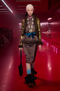 Model walking a neon‑pink runway in a gold sequin coat, fur trim, and patterned dress with a blue bow and long beaded necklace.
