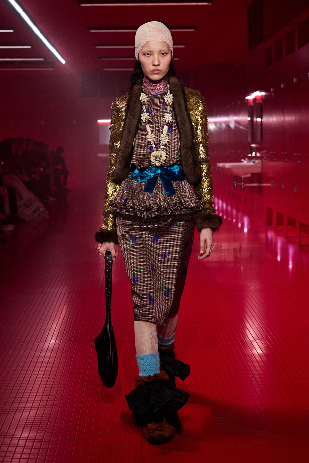 Model walking a neon‑pink runway in a gold sequin coat, fur trim, and patterned dress with a blue bow and long beaded necklace.