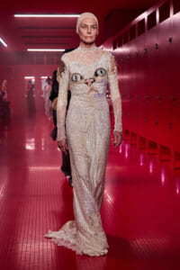 Model walking down a red-lit backstage hallway in a full-length ivory sequin lace gown with a cat face design on the bodice and a matching headpiece.
