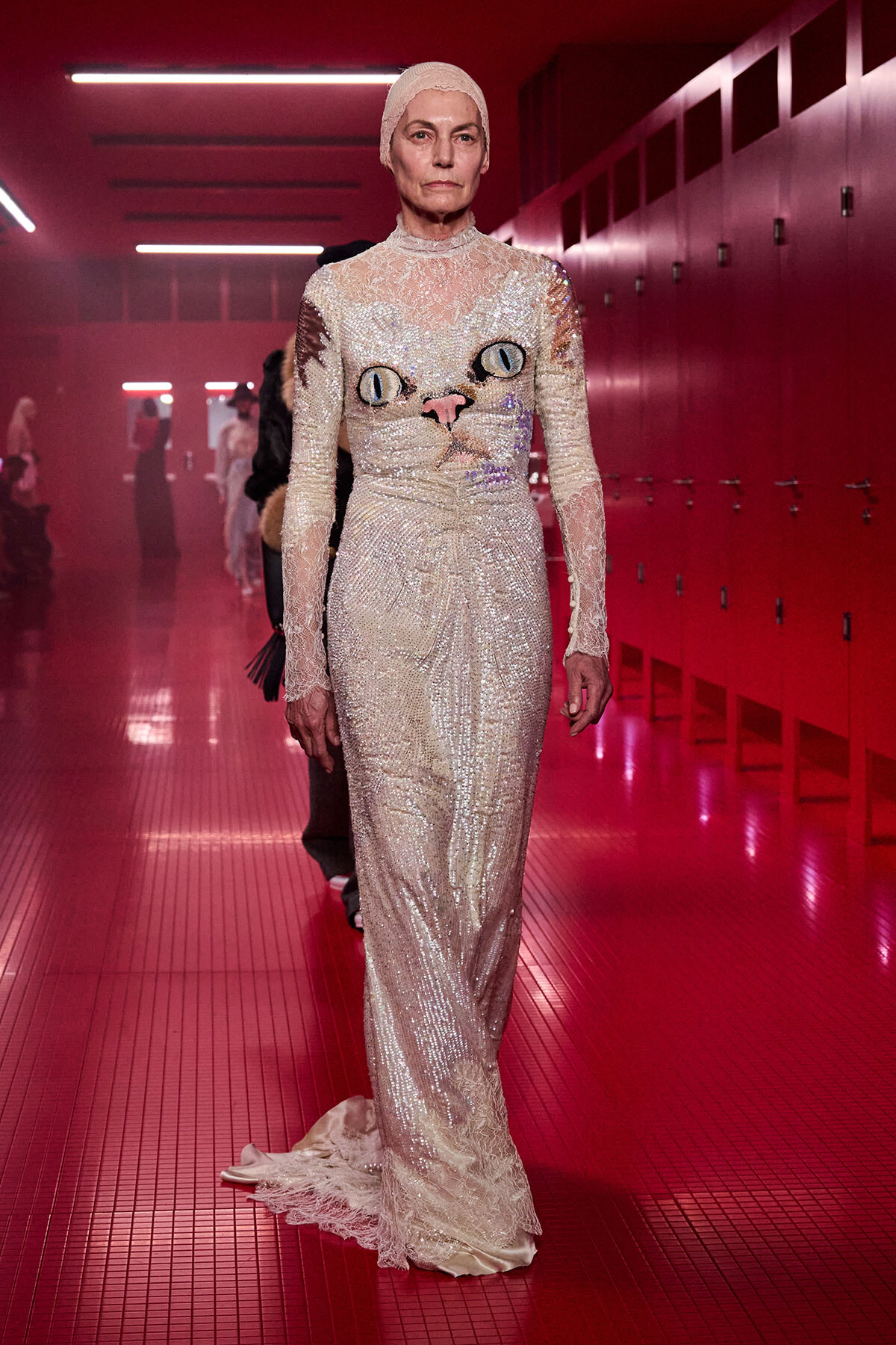 Model walking down a red-lit backstage hallway in a full-length ivory sequin lace gown with a cat face design on the bodice and a matching headpiece.