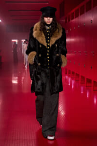 Model walking in a long black fur coat with tan fur trim, black cap, and polka-dot tie in a red-lit hallway.