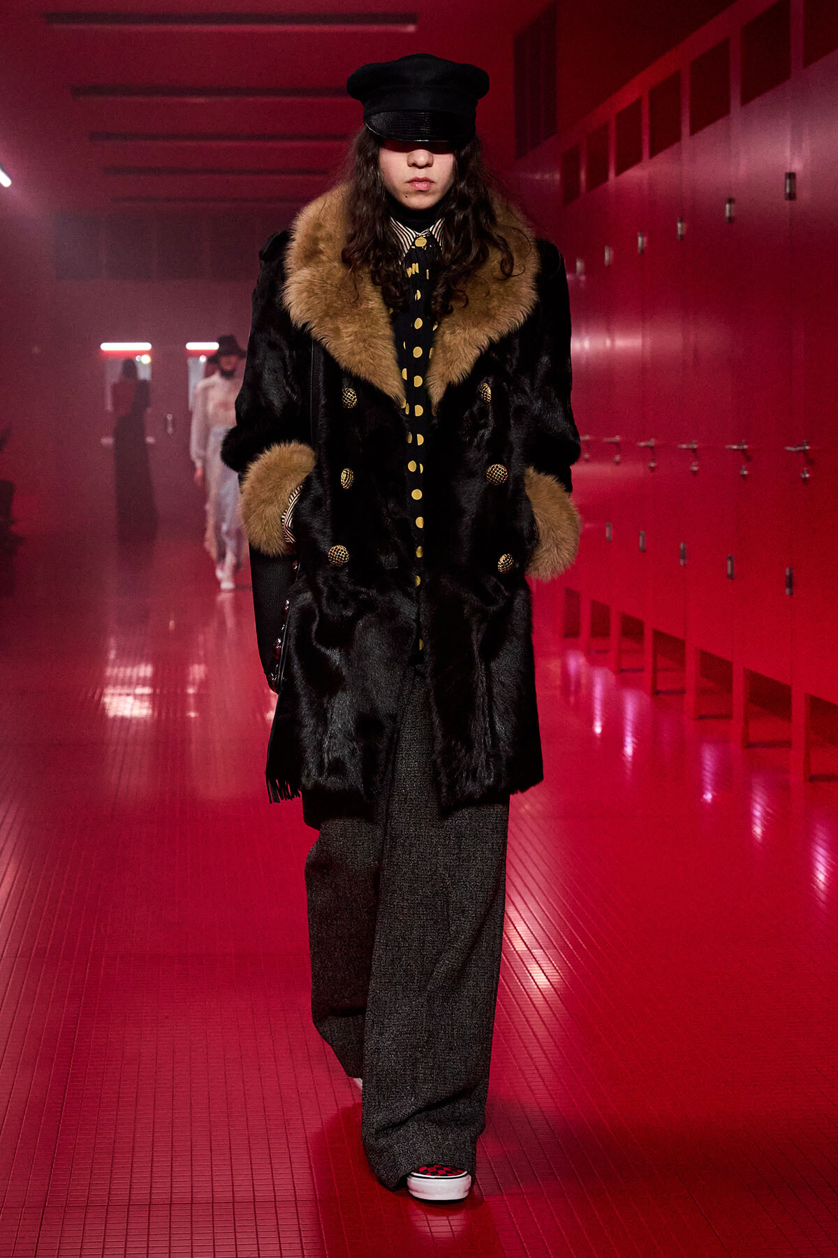 Model walking in a long black fur coat with tan fur trim, black cap, and polka-dot tie in a red-lit hallway.