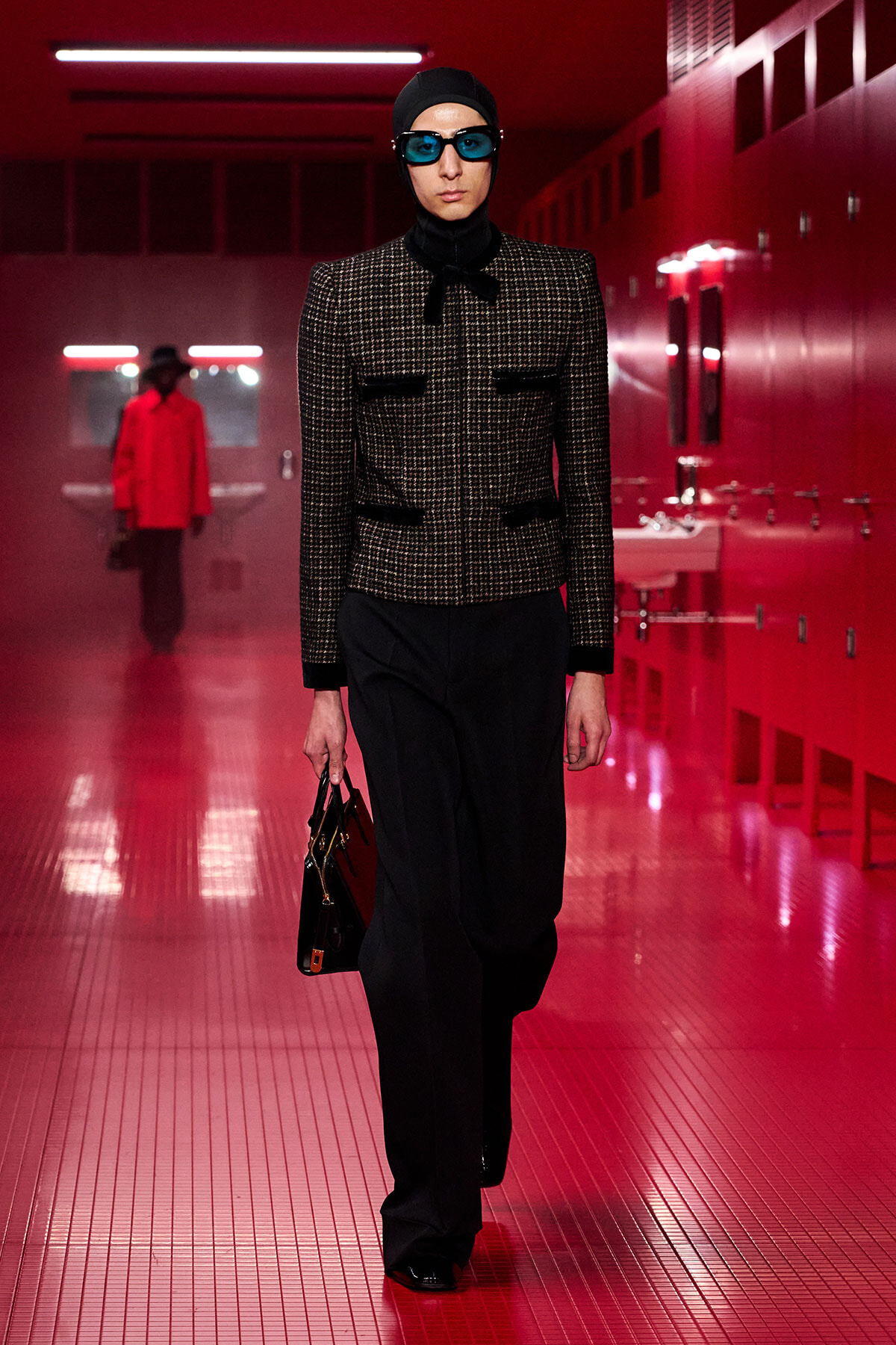 Model walking a runway in a red-lit locker room, wearing a tweed jacket, black hijab, blue sunglasses, wide-leg pants, and carrying a handbag.