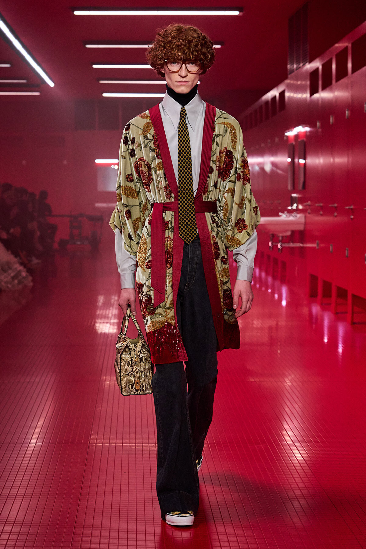 Male runway model walks down a pink-lit hallway in a floral kimono-style robe over a white shirt, yellow dotted tie, and dark wide-leg jeans, carrying a snakeskin handbag.