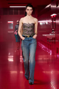 Model walking a runway in a strapless gray corset with lace and bow accents, paired with blue jeans in a red-lit corridor.
