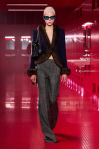 Model wearing a navy fur-trim blazer with fur cuffs and gray wide-leg pants, beige cap, blue sunglasses, and a black chain-strap bag in a red-lit hallway.