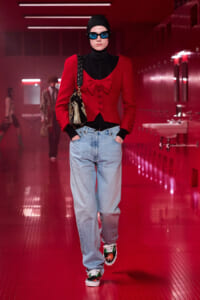 Model in a red cardigan over a black turtleneck, bag on shoulder, light-wash jeans, colorful sneakers, blue sunglasses in a red-lit hallway.