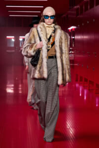 Model walking a runway in a beige fur coat, oversized gray trousers, tan top, beige headscarf, blue sunglasses, and a black handbag in a red-lit hallway.