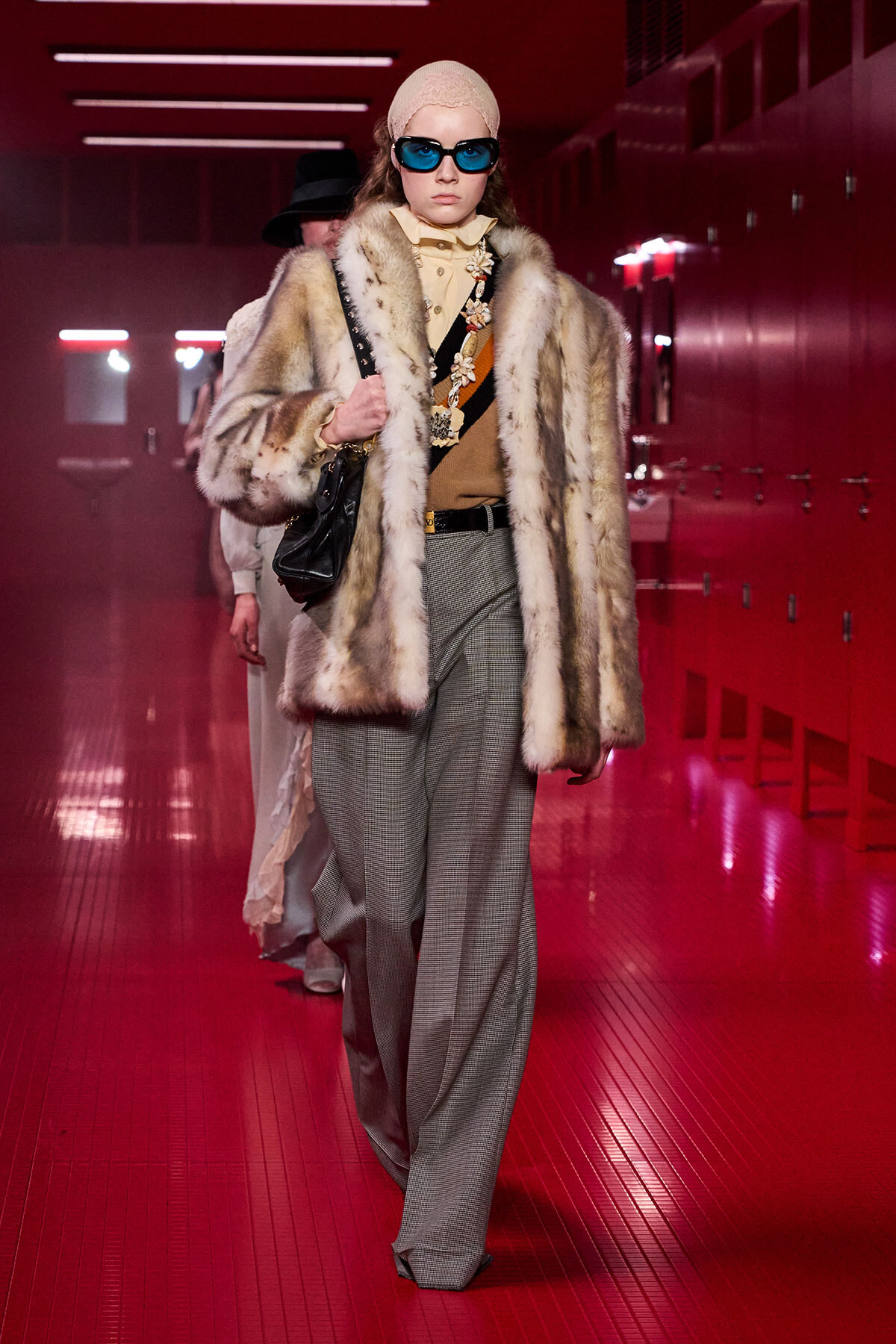 Model walking a runway in a beige fur coat, oversized gray trousers, tan top, beige headscarf, blue sunglasses, and a black handbag in a red-lit hallway.