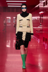 Model on a pink-lit runway wearing a cream textured suit with black trim, belt, and black fringe details, plus bright green knee-high socks and blue sunglasses.