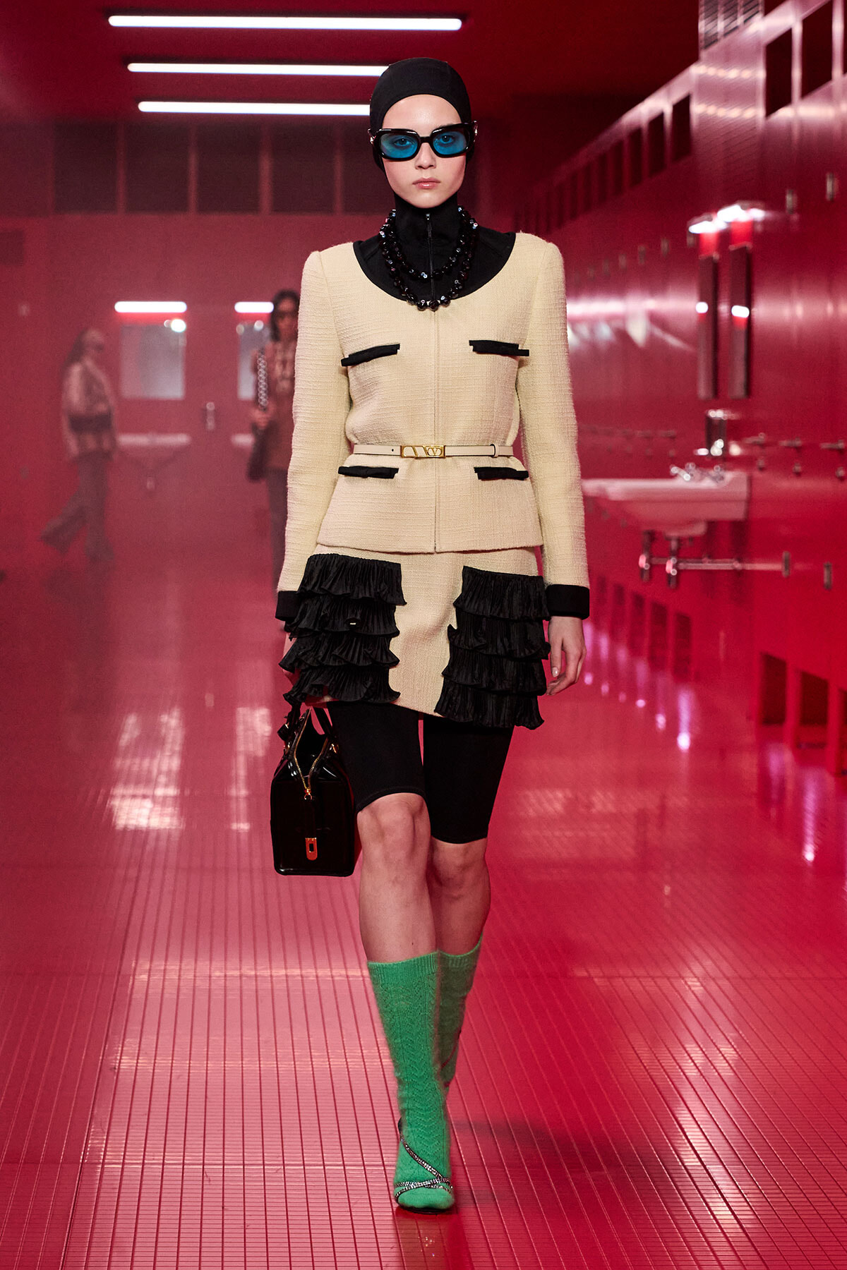 Model on a pink-lit runway wearing a cream textured suit with black trim, belt, and black fringe details, plus bright green knee-high socks and blue sunglasses.