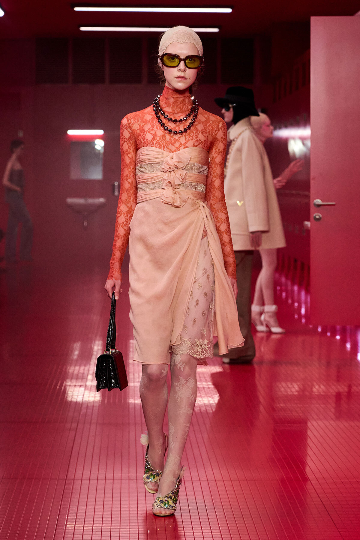 Model in peach lace high-neck dress with a wrapped sash and floral detail, walking a pink-lit runway wearing yellow sunglasses and a beige head wrap, carrying a small black bag.