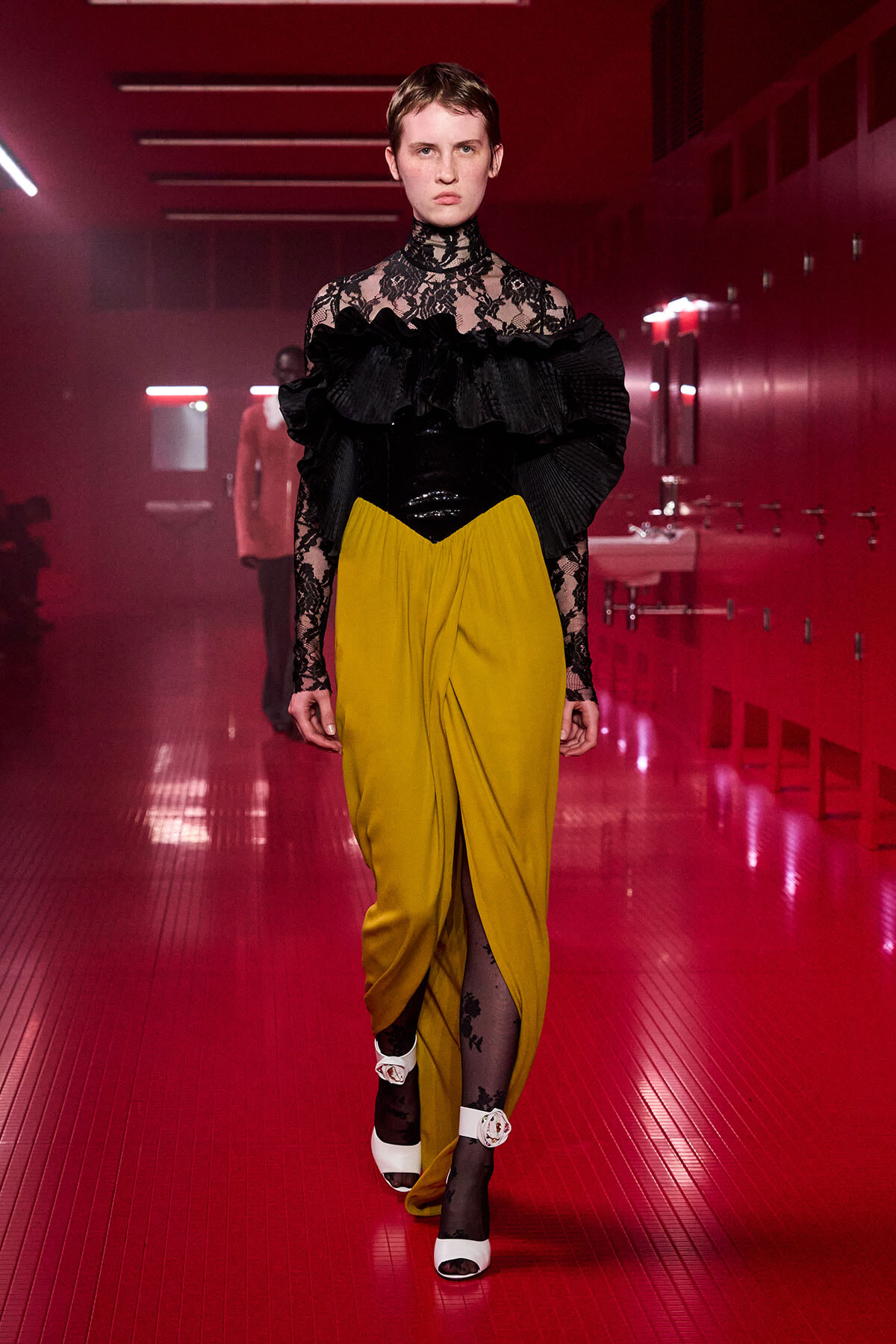 Model walking down a red-lit hallway in a black lace high-neck top with oversized ruffles and mustard wide-leg pants, white platform sandals with ankle straps.
