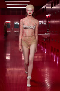 Model walking a runway in pale pink lace bodysuit with satin bust, beige head cap, and white heels in a red-tiled backstage area.