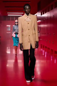 Model wearing a beige tailored blazer with a patterned bow necktie walks a red-lit runway with a second model in the background.