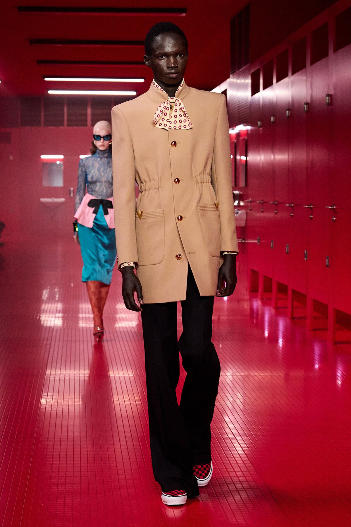 Model wearing a beige tailored blazer with a patterned bow necktie walks a red-lit runway with a second model in the background.