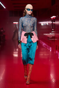 Model walking a red-lit runway in a blue lace turtleneck, pink peplum with a black bow, and teal satin pants, wearing blue sunglasses and a beige headwrap.