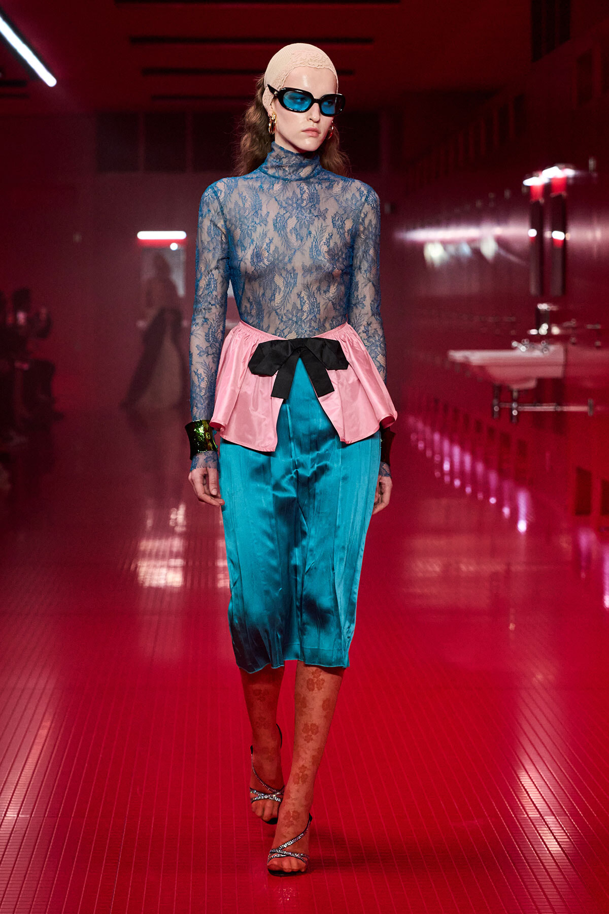 Model walking a red-lit runway in a blue lace turtleneck, pink peplum with a black bow, and teal satin pants, wearing blue sunglasses and a beige headwrap.