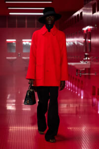 Model wearing a bright red oversized coat with a black hat, black pants, leopard-print shoes, and a black handbag in a red-lit hallway.