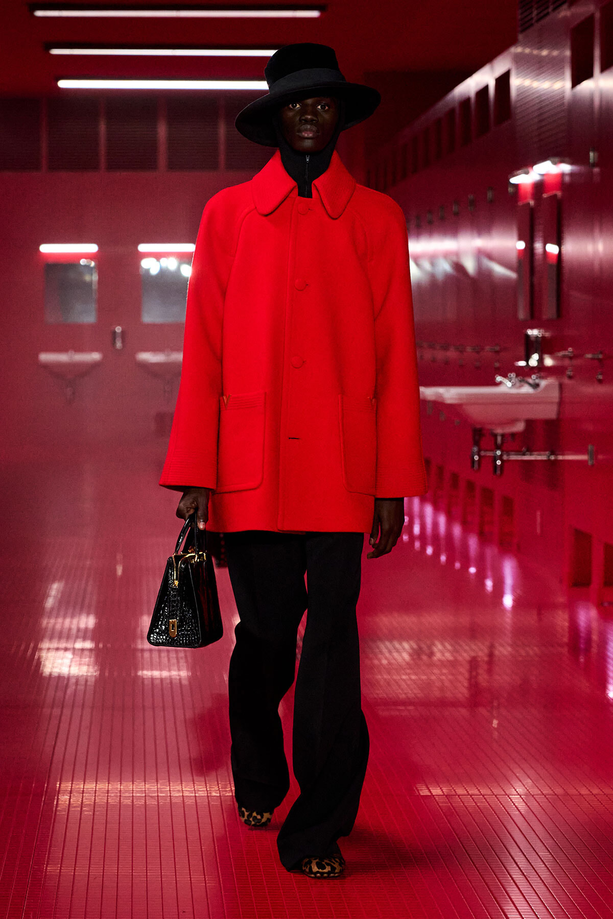 Model wearing a bright red oversized coat with a black hat, black pants, leopard-print shoes, and a black handbag in a red-lit hallway.