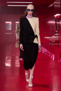 Model on a runway wearing a black-and-cream color-block blazer dress with a waist knot and bold accessories.",