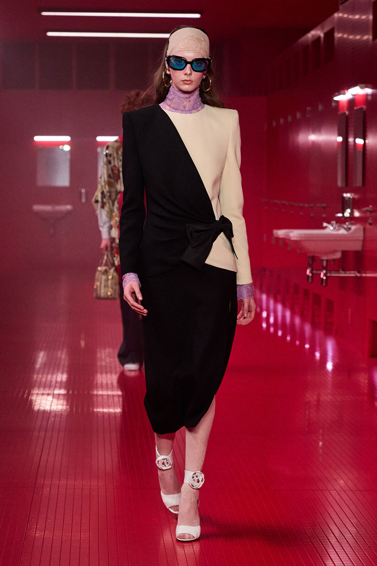 Model on a runway wearing a black-and-cream color-block blazer dress with a waist knot and bold accessories.",