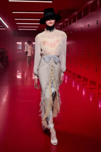 Model walks a fashion runway in a vintage cream lace blouse with sheer bodice and layered ruffle skirt, wearing a wide-brim hat in a red-lit hallway.