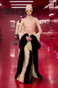 Model walks a fashion show in a sheer nude mesh top, cream wide-leg pants, and a black fur-trimmed cape, wearing pale headscarf and yellow sunglasses in a red-lit backstage hallway.