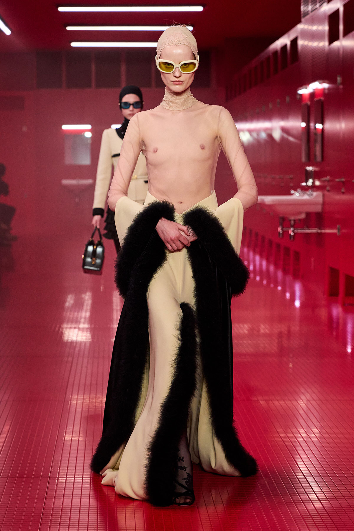 Model walks a fashion show in a sheer nude mesh top, cream wide-leg pants, and a black fur-trimmed cape, wearing pale headscarf and yellow sunglasses in a red-lit backstage hallway.