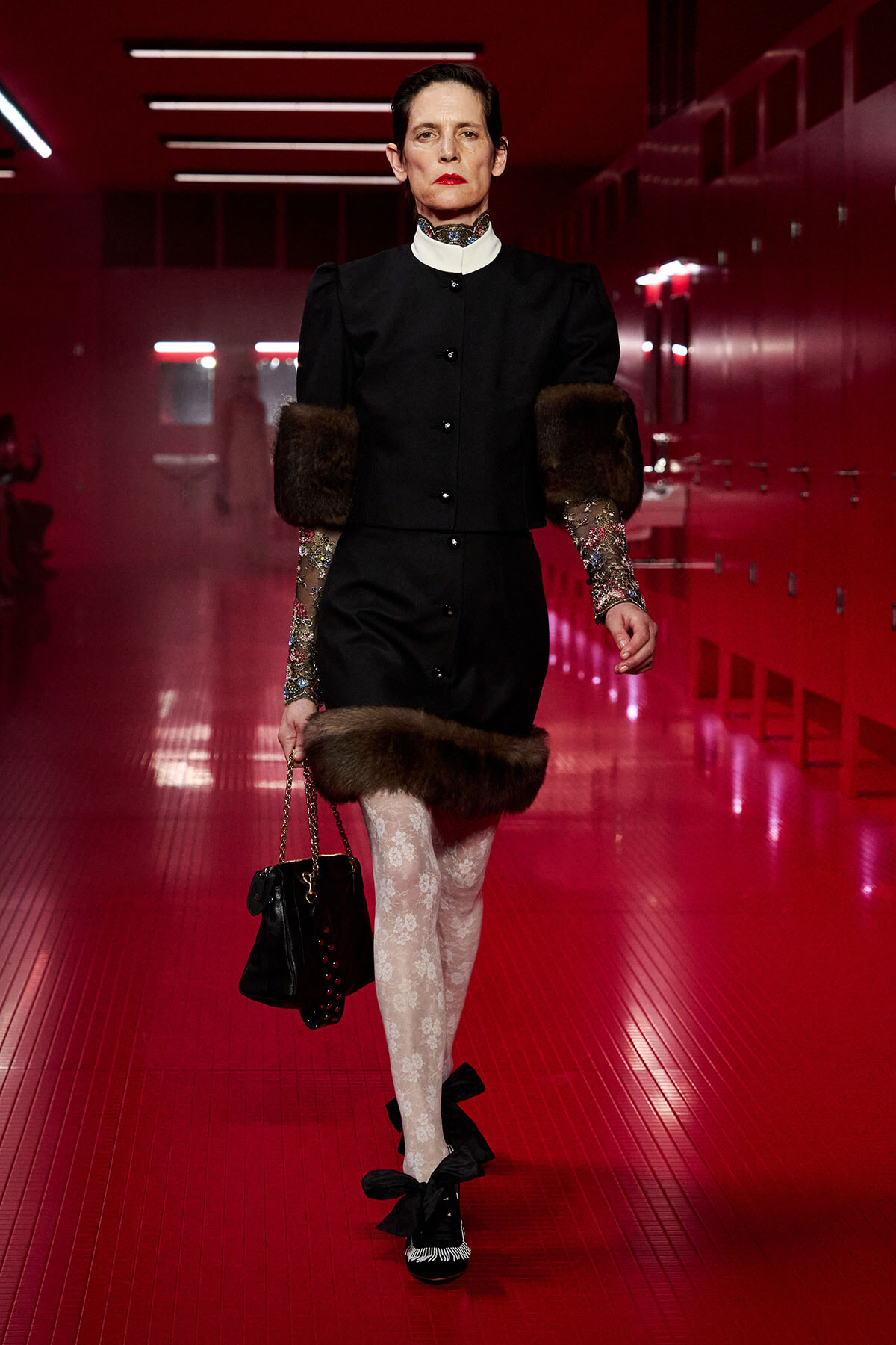 Model on a red-lit runway wearing a black buttoned dress with fur cuffs and hem, patterned sheer sleeves, white lace tights, and black bow-strap heels with a black handbag.