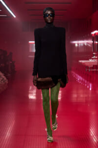 Model walking a fashion show runway in a black turtleneck dress with fur trim at the hem, green patterned tights, embellished heels, and oversized sunglasses under red lighting