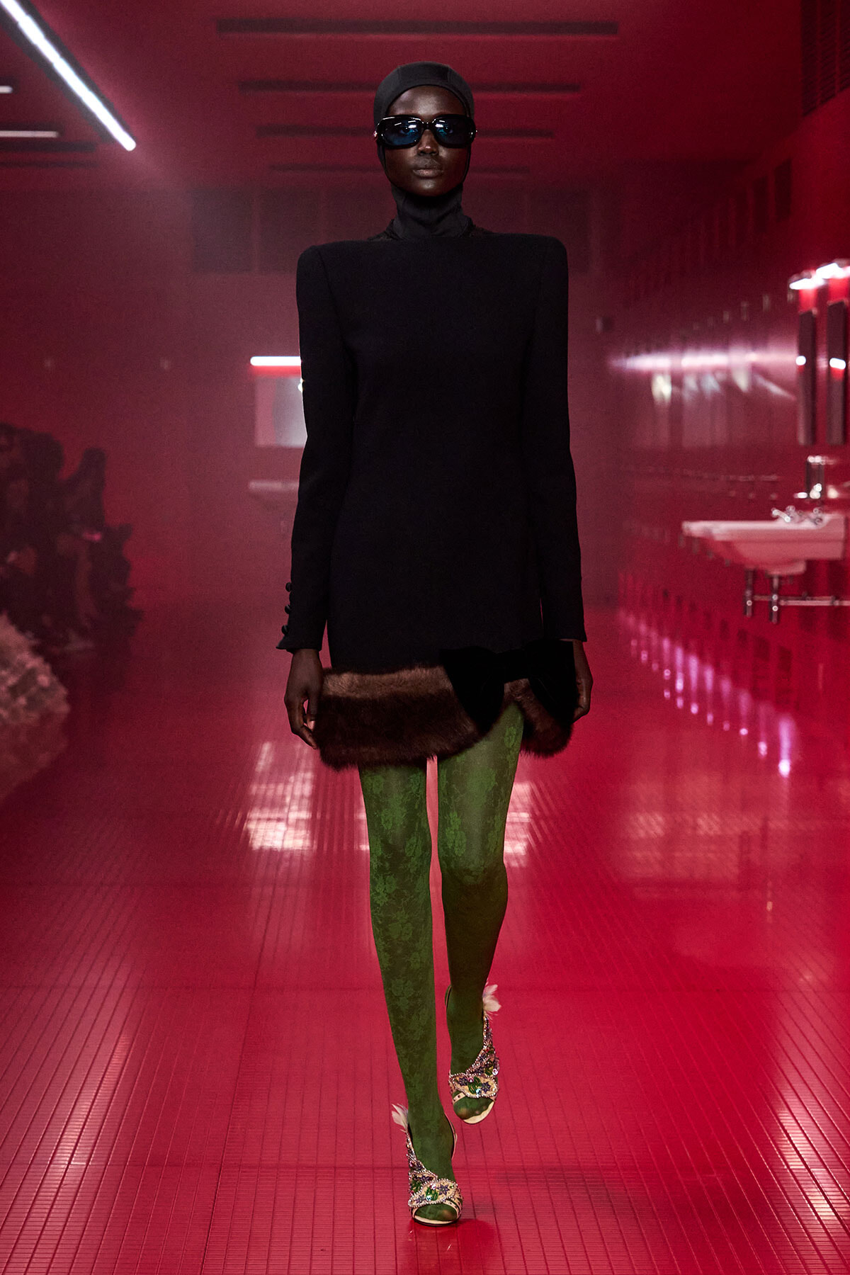 Model walking a fashion show runway in a black turtleneck dress with fur trim at the hem, green patterned tights, embellished heels, and oversized sunglasses under red lighting