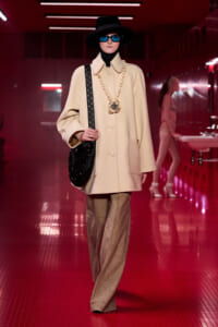 Model on a red-lit runway wearing a cream oversized coat, black wide-brim hat, blue sunglasses, chunky gold necklace, and a black quilted shoulder bag.