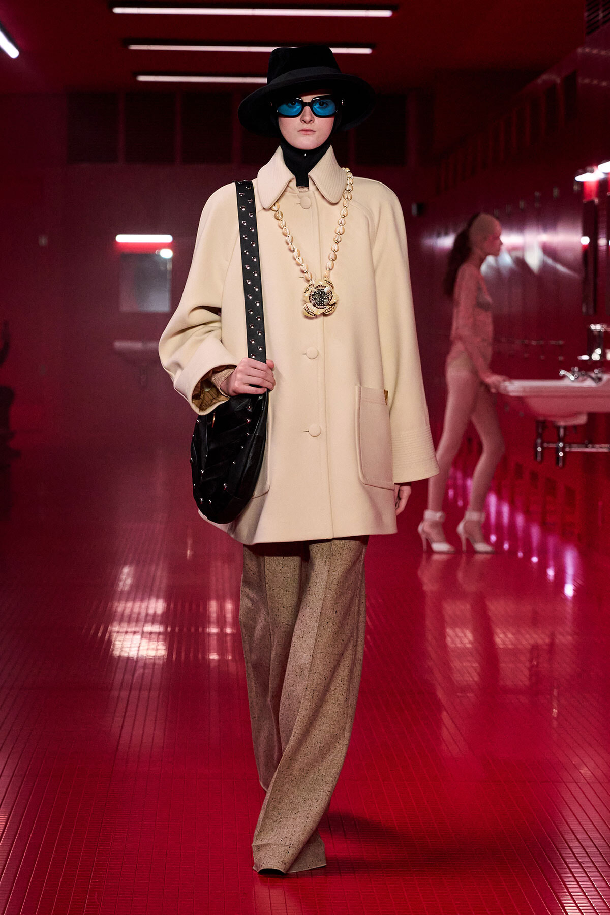 Model on a red-lit runway wearing a cream oversized coat, black wide-brim hat, blue sunglasses, chunky gold necklace, and a black quilted shoulder bag.