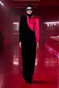 Model walks a red-lit runway in a two-tone velvet gown: black on one side, magenta sash on the other, with a head wrap and blue sunglasses.
