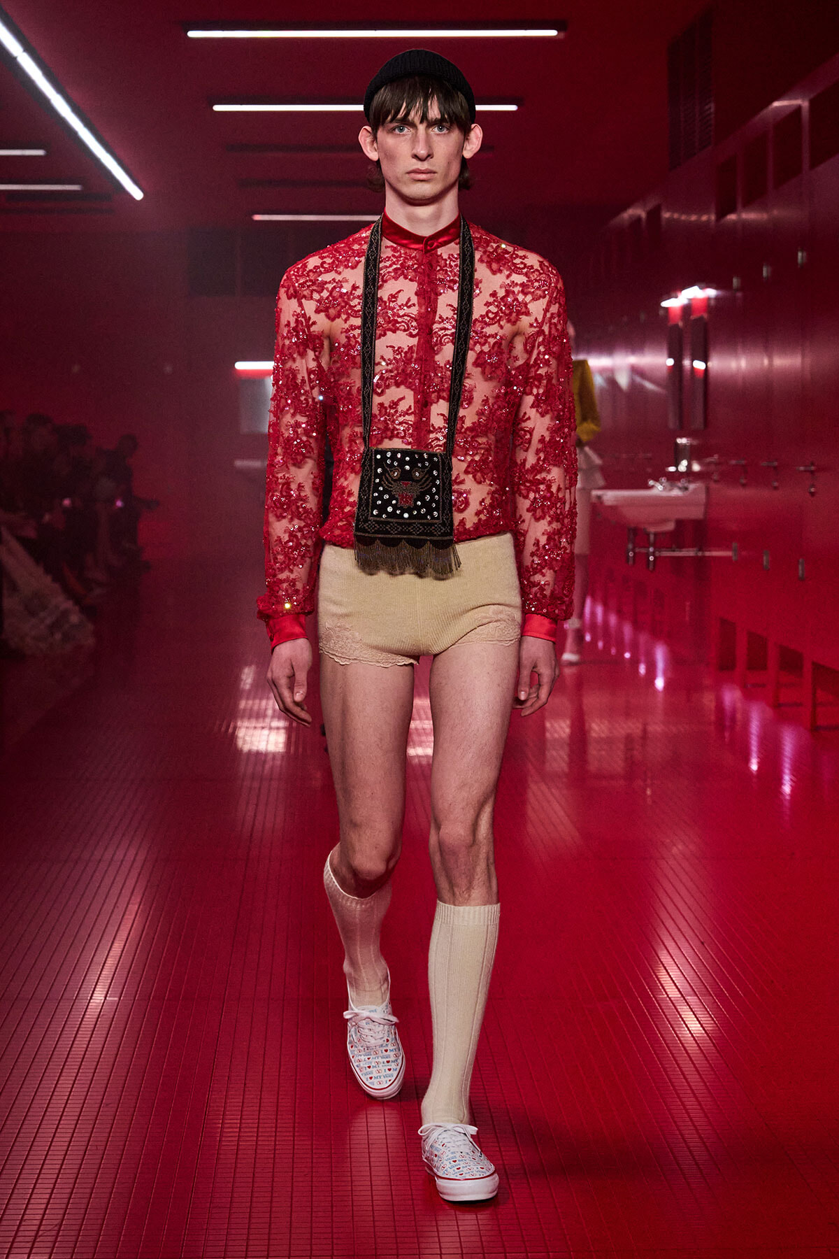 Male model walking a runway in a red lace sheer top, beige lace shorts, cream knee-high socks, white sneakers, a black crossbody bag, and a black beanie.
