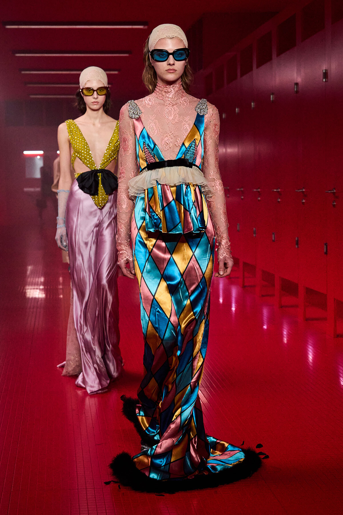 Model walking a runway in a bold multicolor harlequin gown with pink lace sleeves and blue sunglasses in a red-lit corridor gala setting.