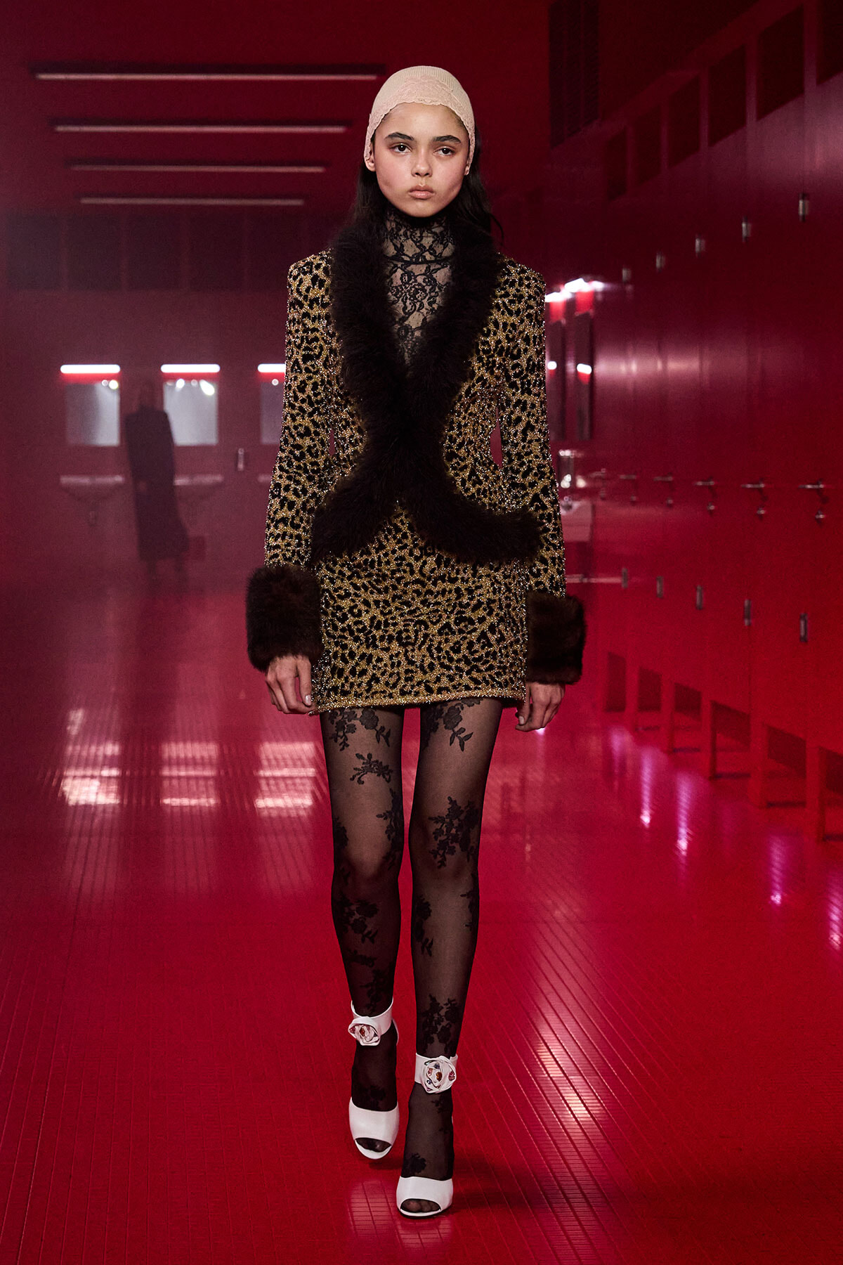 Model wearing a leopard-print coat with black fur trim and lace top, walking in a red-lit locker-room hallway