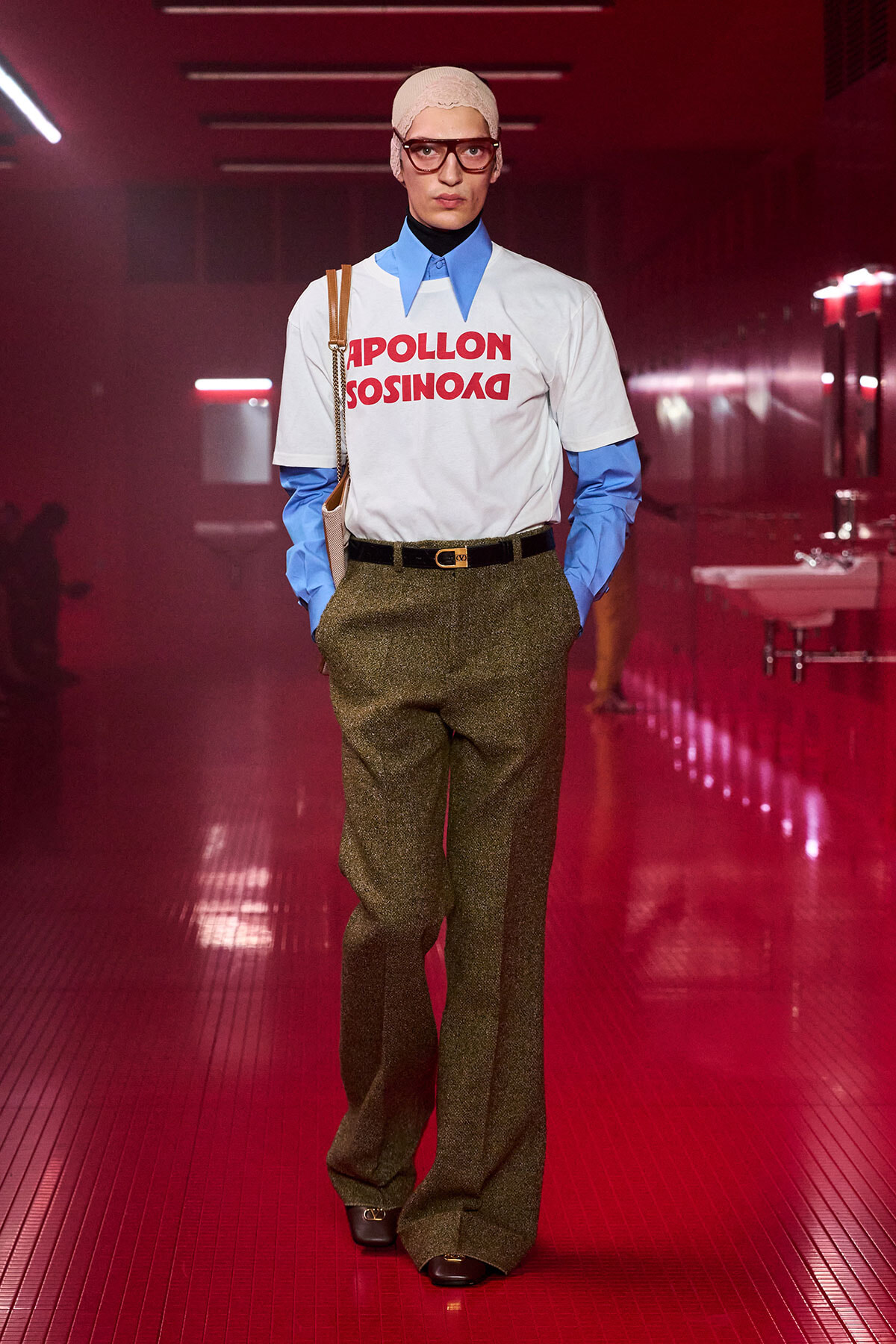 Runway shot of a model in a retro-inspired outfit: white T‑shirt with red text over a blue shirt, glittery olive trousers, black belt, beige head wrap, glasses, and a shoulder bag in a pink-lit hallway.