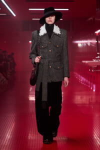 Model walks a red-lit runway wearing a gray tweed blazer with gold buttons, a white ruffled collar, black wide-brim hat, and black wide trousers.