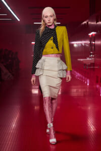 Model walking a runway in a bold color-block outfit: lime-yellow blazer with a black polka-dot sleeve, pink lace underlayers, a cream tiered peplum skirt, and pink lace tights with white heels.