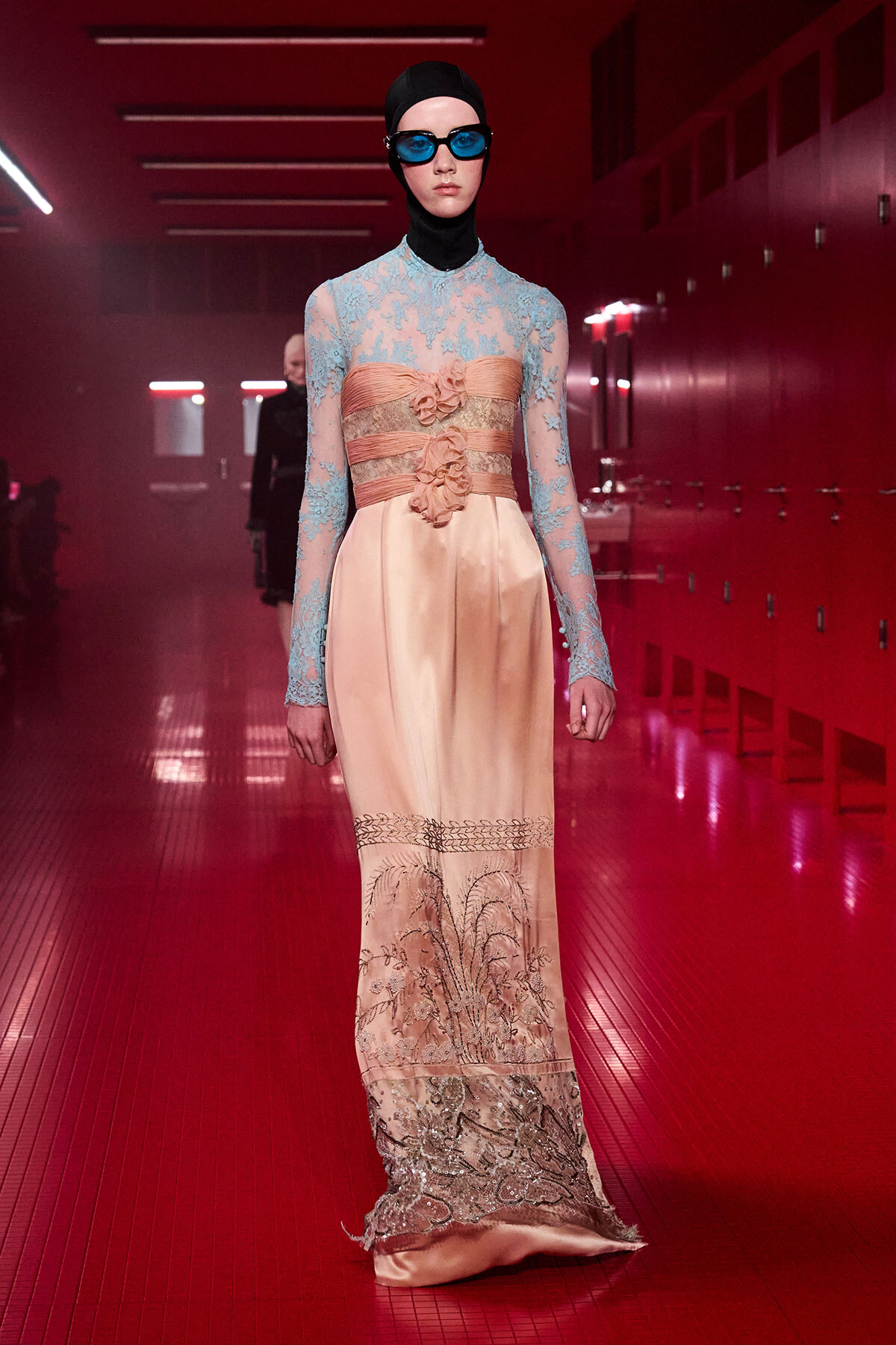 Runway model in a peach satin gown with blue lace bodice and waist corset-style belt, headscarf, and blue goggles in a red-lit backstage area.