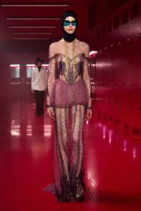 Model on a red-lit fashion runway wearing a sheer pink gown with black bead embellishments, a black head covering, blue sunglasses, and long blonde braids.