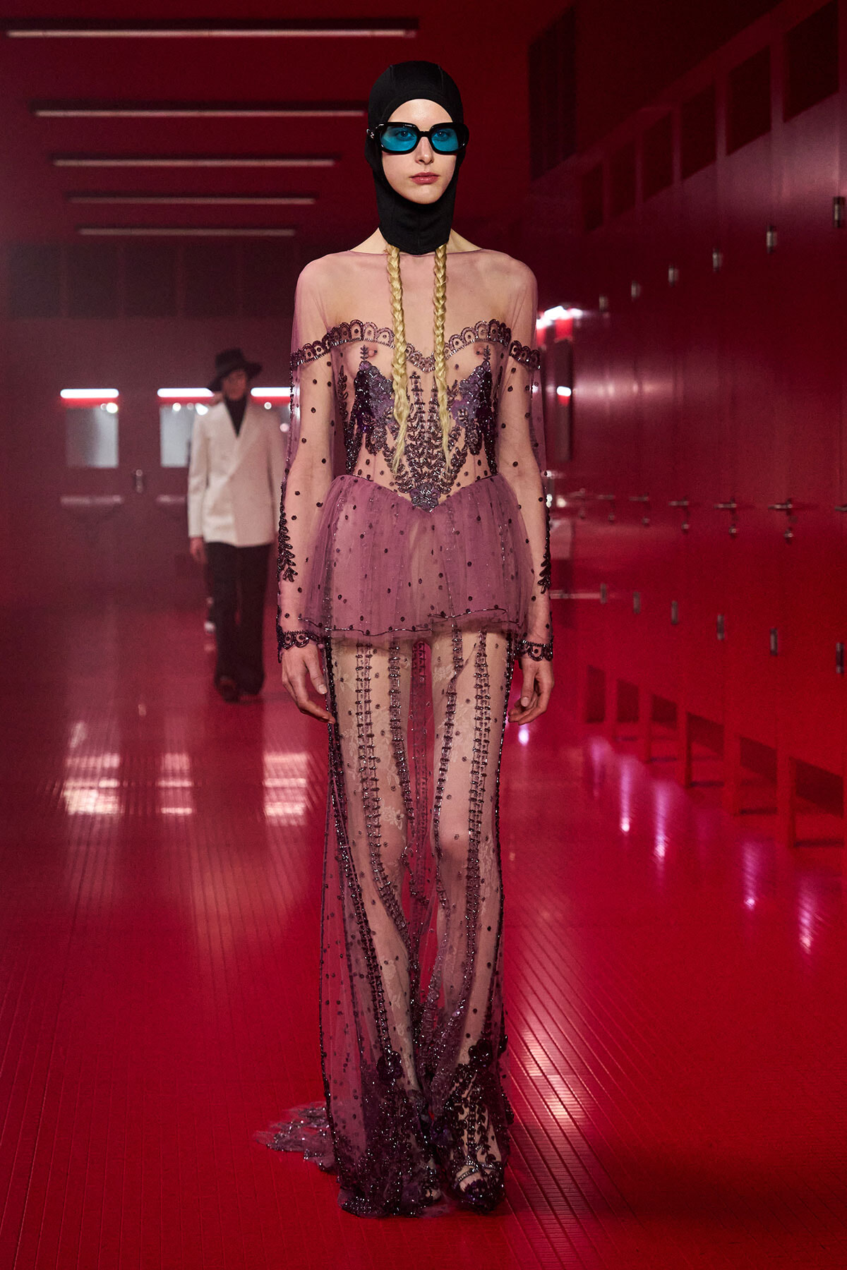 Model on a red-lit fashion runway wearing a sheer pink gown with black bead embellishments, a black head covering, blue sunglasses, and long blonde braids.
