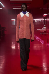 Model walking a fashion runway in a pink knit cardigan with a ruffled white collar and a blue turtleneck, dark wide-leg pants, in a pink-red-lit hallway setting.