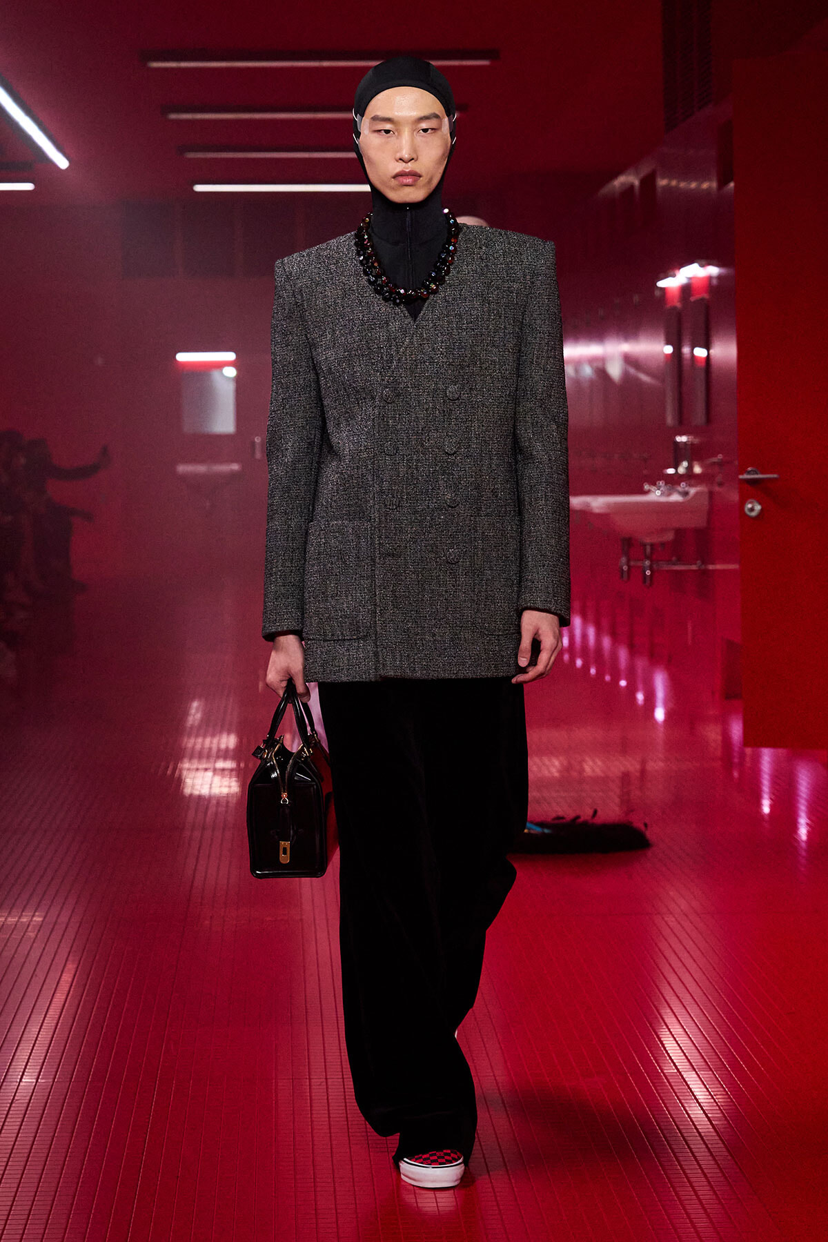 Model walking a red-lit runway in a gray double-breasted blazer, black turtleneck, head covering, and black handbag.