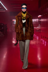 Model walking a fashion runway in a brown fur coat, yellow blouse, gray trousers, black headwrap and sunglasses under red lighting.