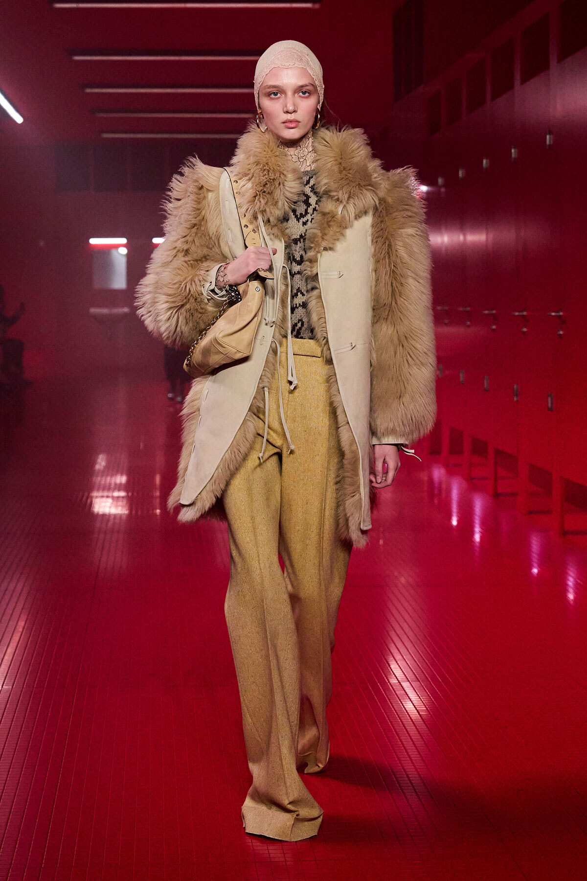Model wearing a beige fur-trimmed coat over a animal-print top, mustard pants, and a knit cap in a red-lit hallway, carrying a tan bag.